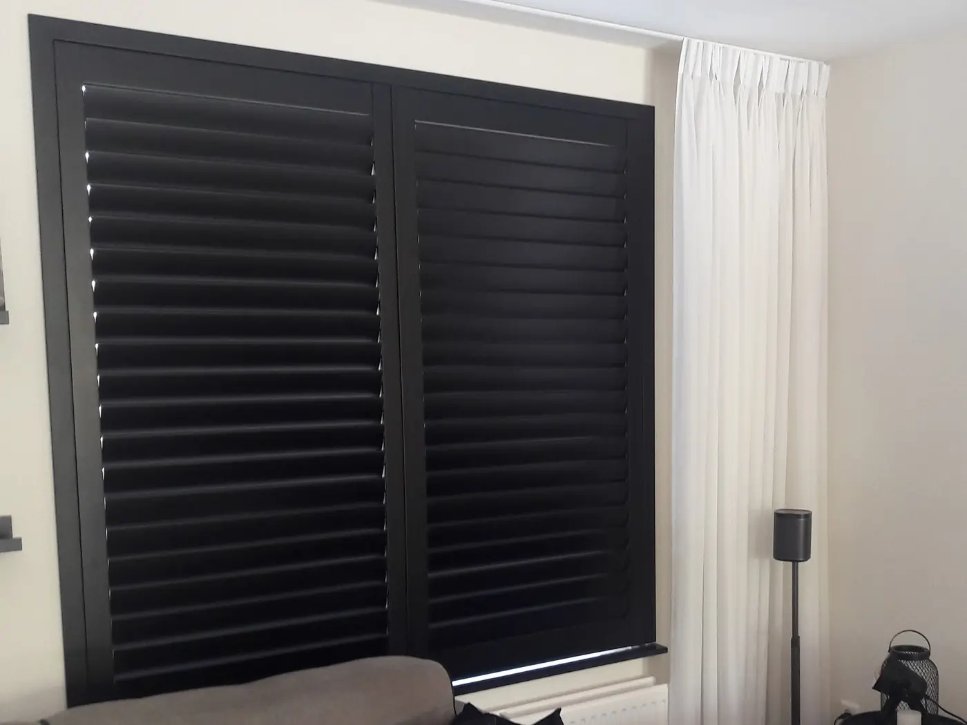 Antracietkleurige shutters voor een woonkamerraam, met witte vitrage ervoor